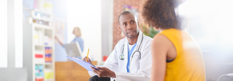A doctor holding a clipboard and consulting a patient seated in a clinic